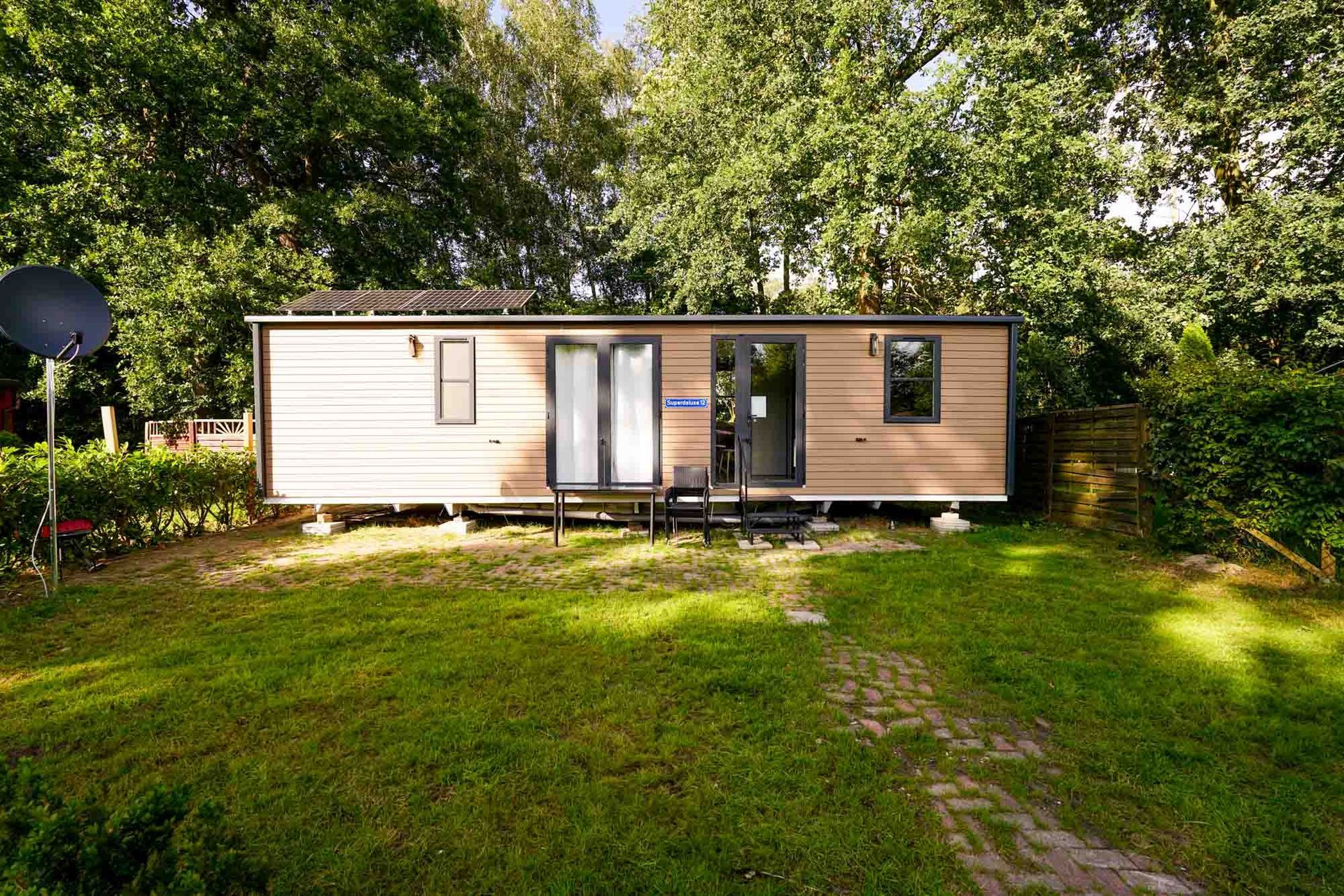 Kleines Mobilheim mit hellbrauner Außenverkleidung, umgeben von grünem Gras und Bäumen.