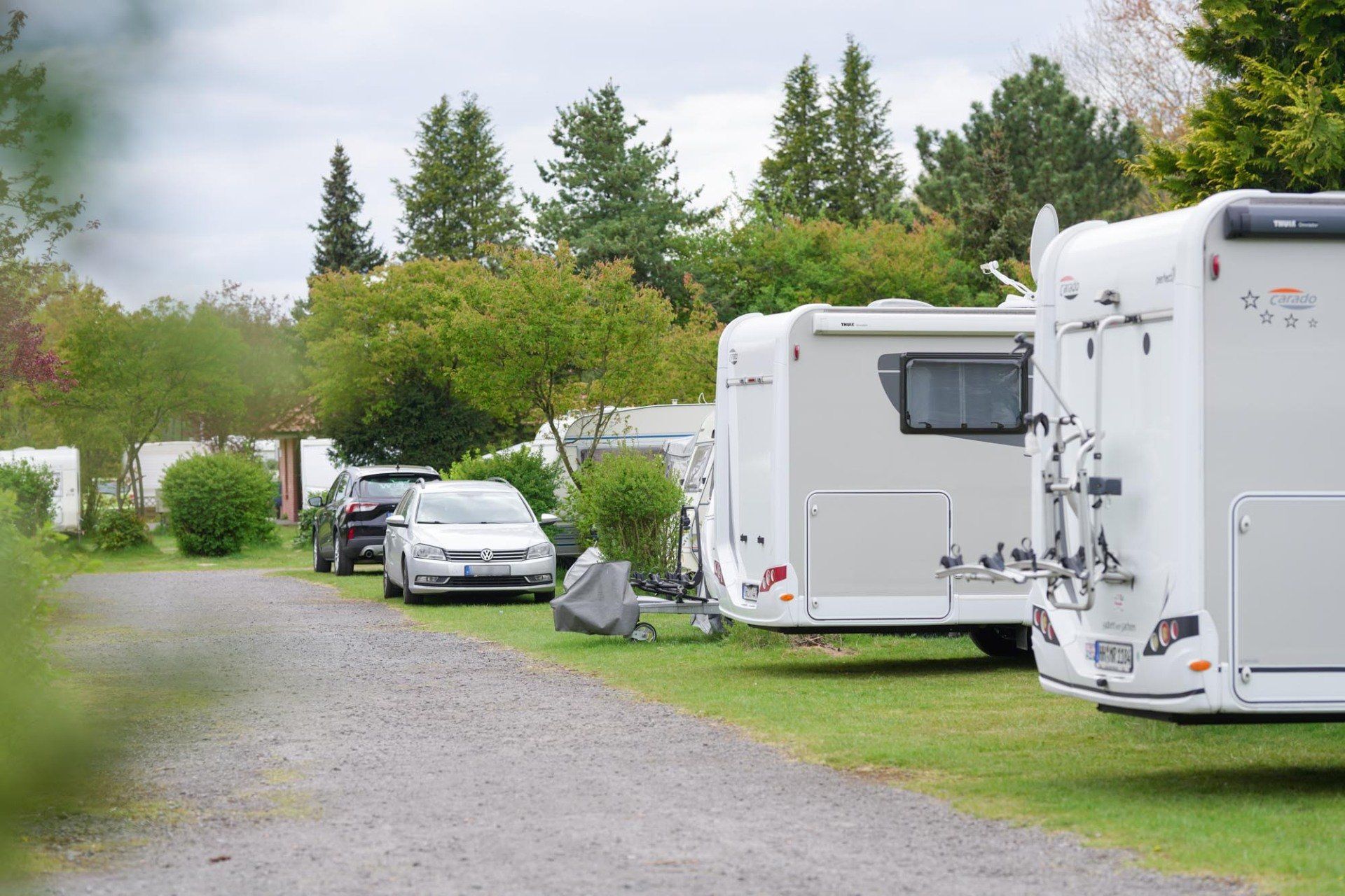 Campingplatz mit Wohnmobilen auf Gras und Schotter, Bäume im Hintergrund