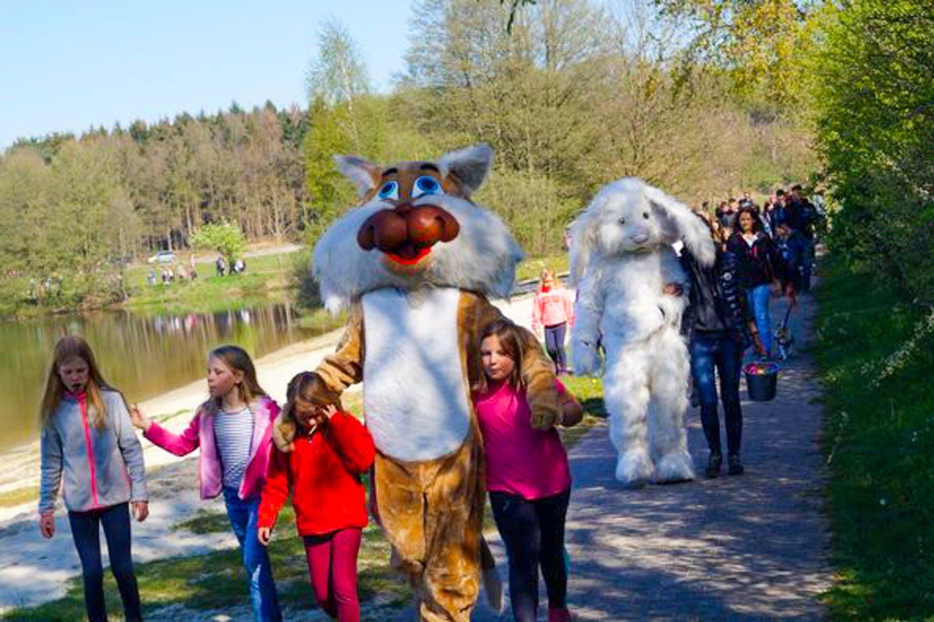 Kinder gehen mit kostümierten Hasen- und Tigerfiguren in der Nähe eines Sees spazieren.