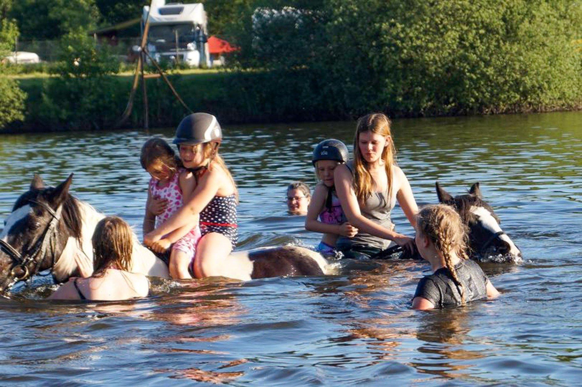 Kinder in Badeanzügen reiten und spielen mit Pferden im Wasser.