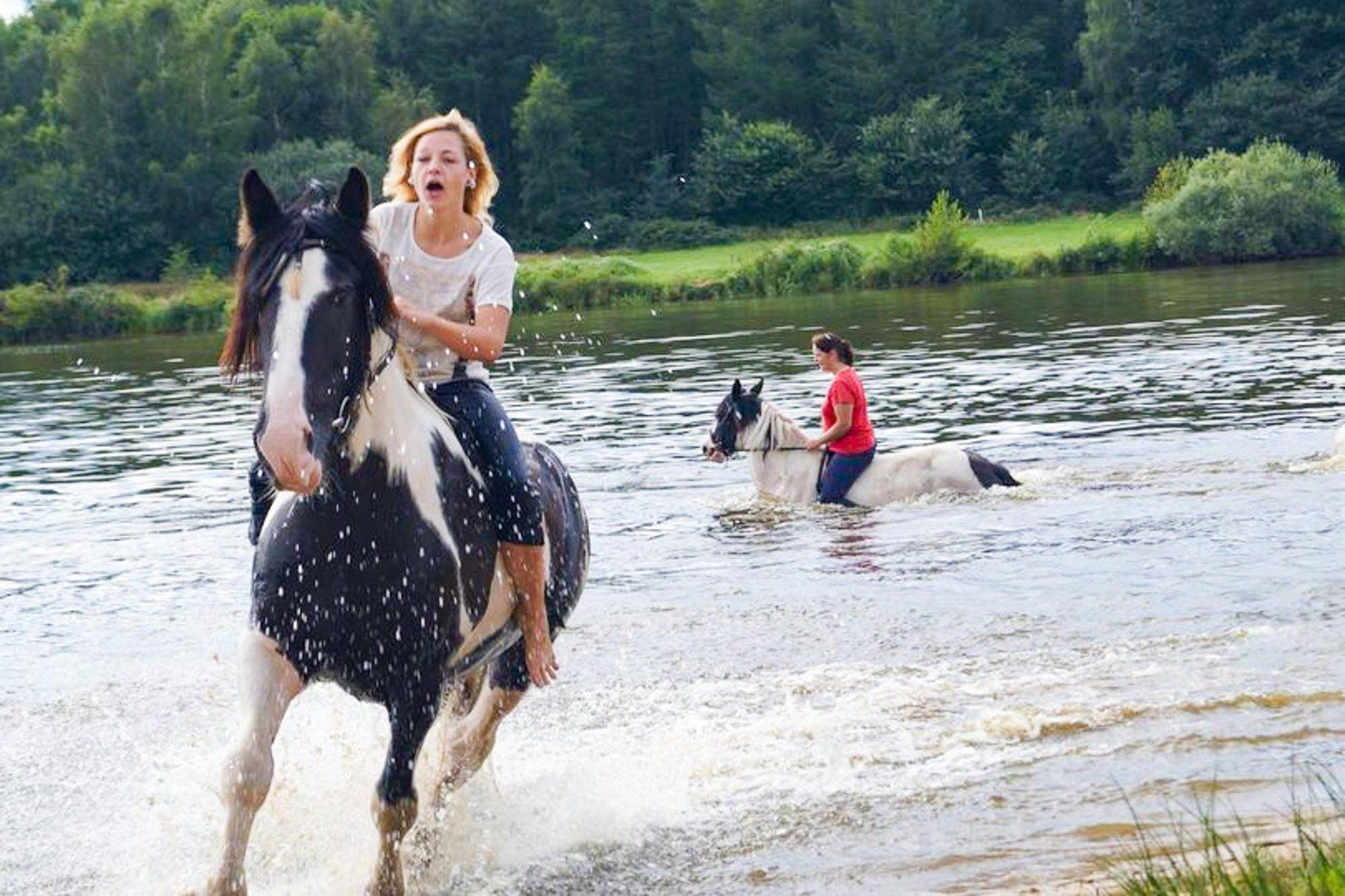 Frau reitet auf schwarz-weißem Pferd durchs Wasser, zweites Pferd im Hintergrund.