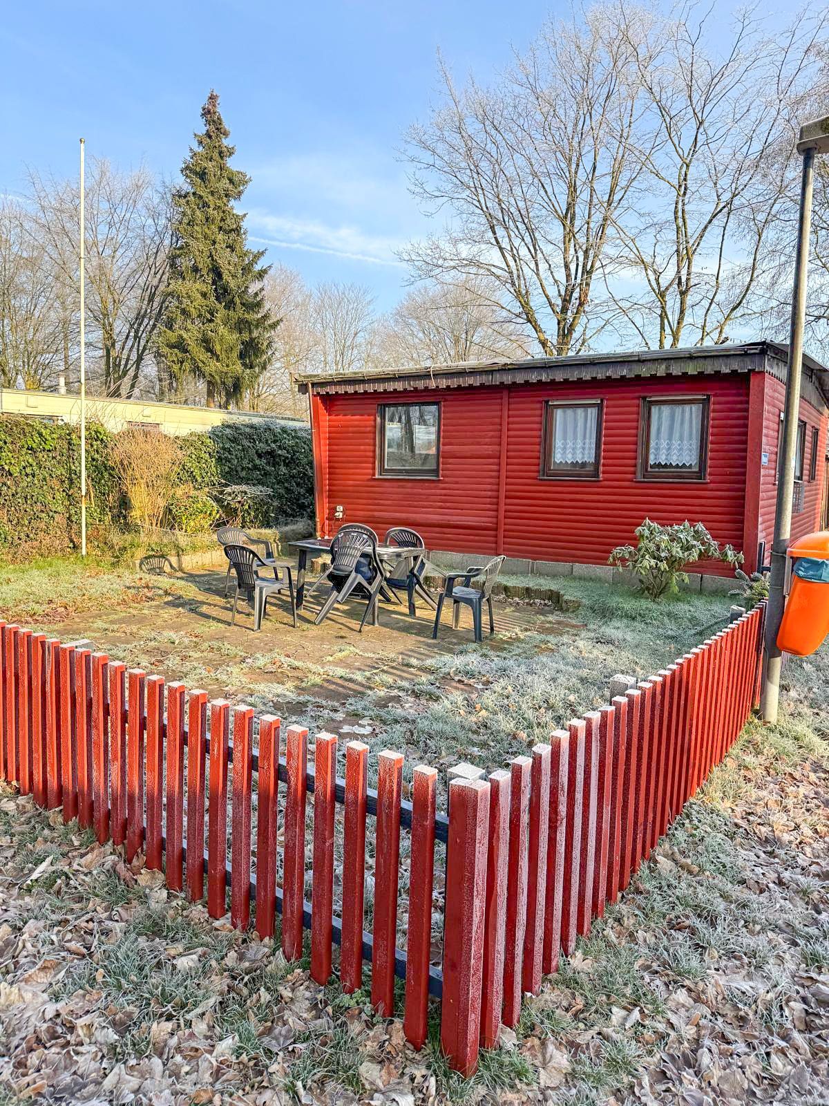 Rote Hütte mit rotem Lattenzaun auf einem Rasengrundstück. Sitzplätze im Freien sichtbar. Kaltes, möglicherweise frostiges Wetter.