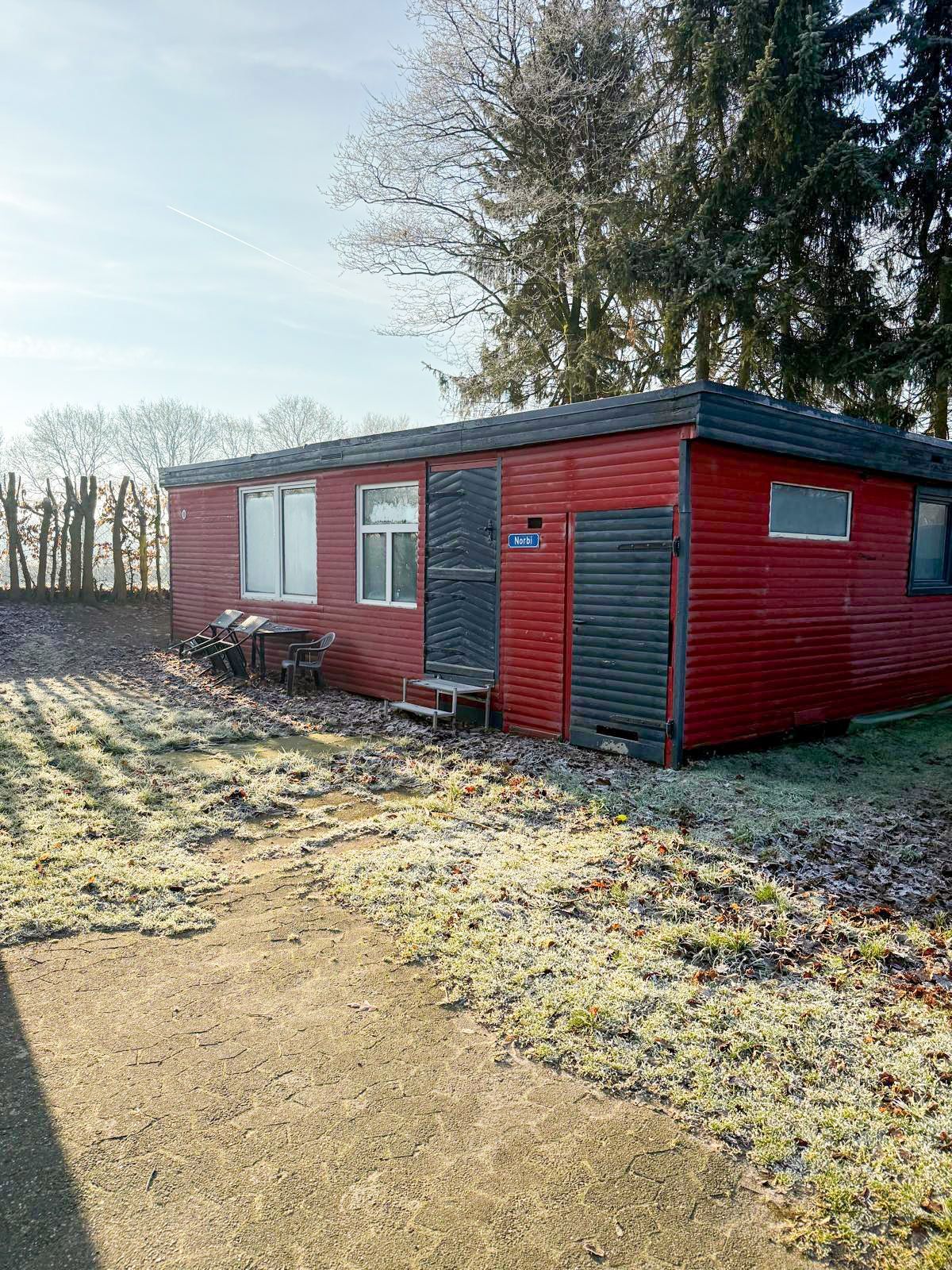Roter Holzschuppen mit schwarzer Tür und Fenster, auf einer Wiese mit Bäumen im Hintergrund.