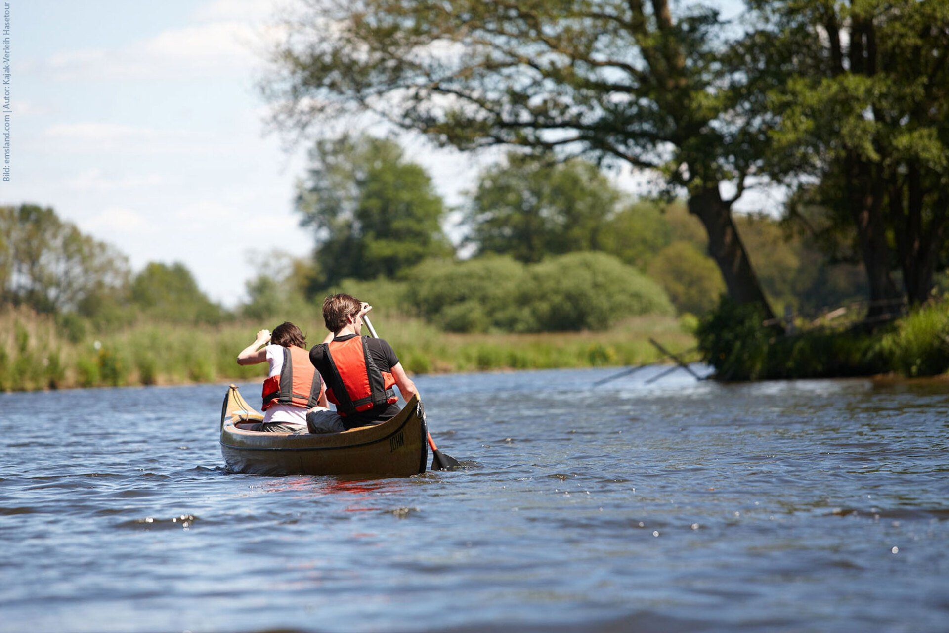 Bild von www.paddel-und-pedal.de Zwei Personen fahren unter blauem Himmel Kanu auf einem Fluss, umgeben von Bäumen und Schilf.