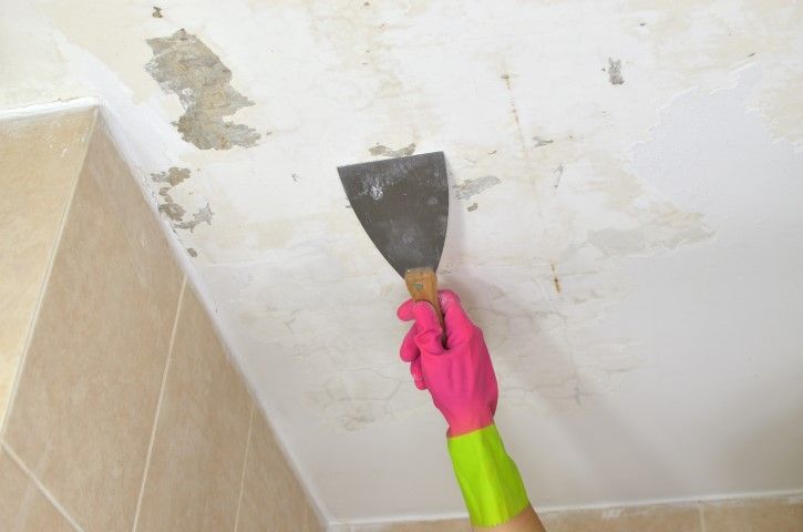 Person scraping peeling paint from a ceiling with a putty knife, wearing a pink and green glove.