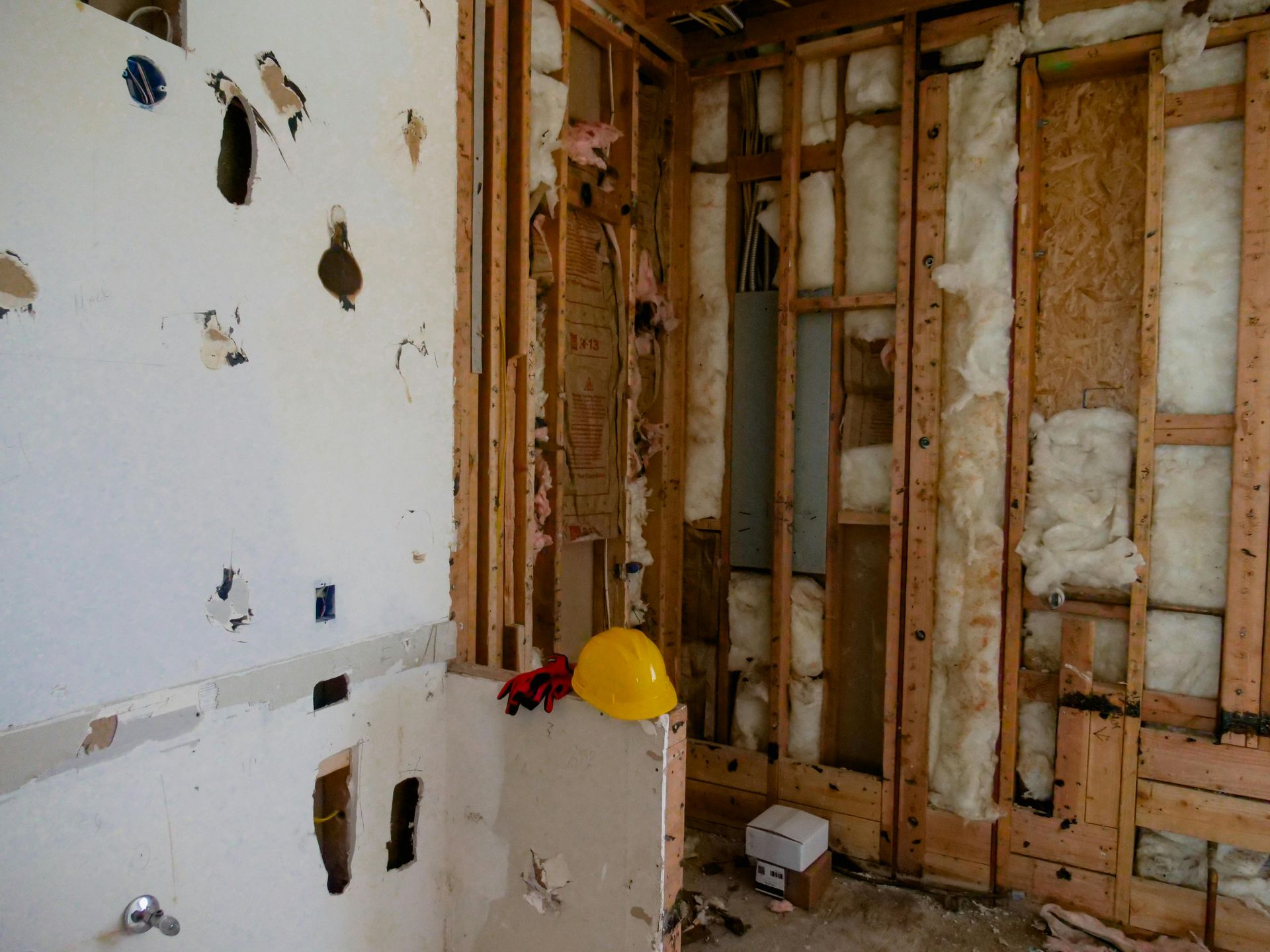 Partially demolished interior wall with exposed studs, insulation, tools, and a yellow hard hat on the floor