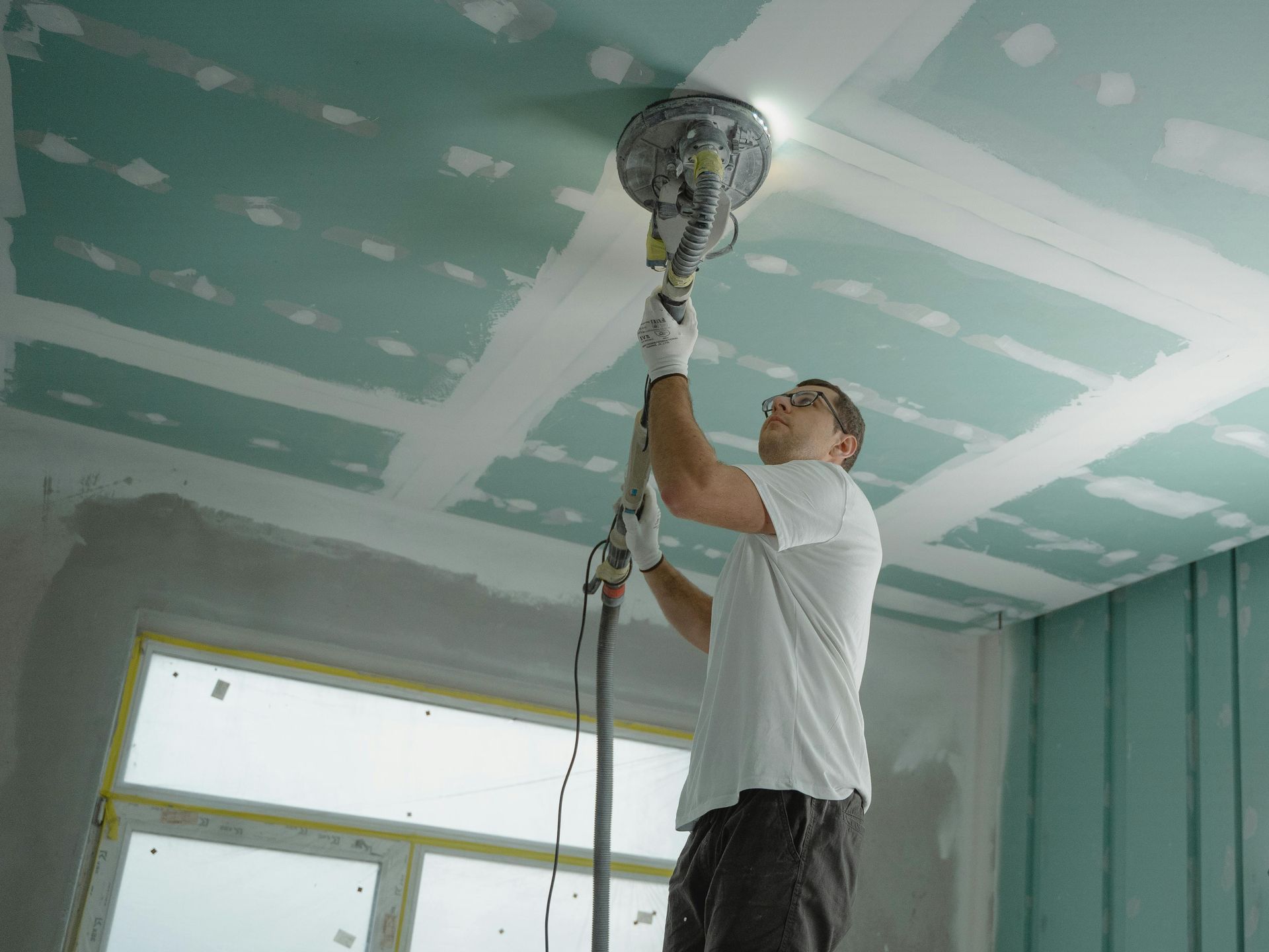 Worker sanding a teal ceiling with a pole sander in a room under renovation