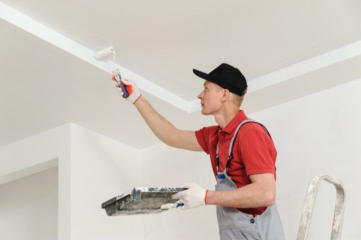Person painting a white ceiling with a paint roller while standing on a ladder.