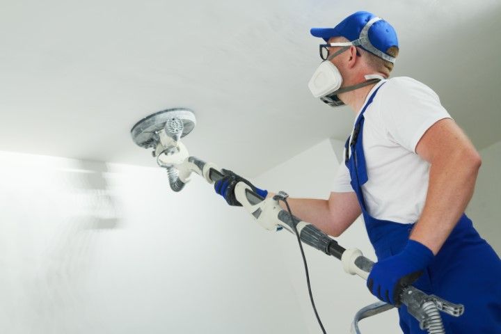 Man sanding a ceiling, wearing a respirator, safety glasses, gloves, and overalls.