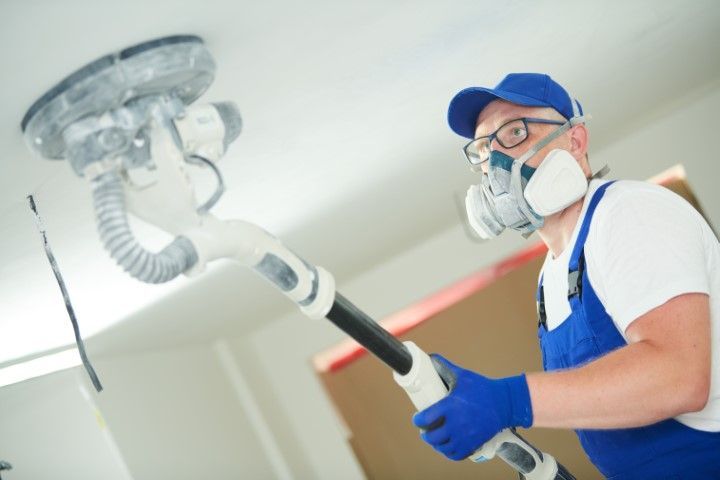 Man sanding a ceiling, wearing a respirator, blue gloves, and hat.