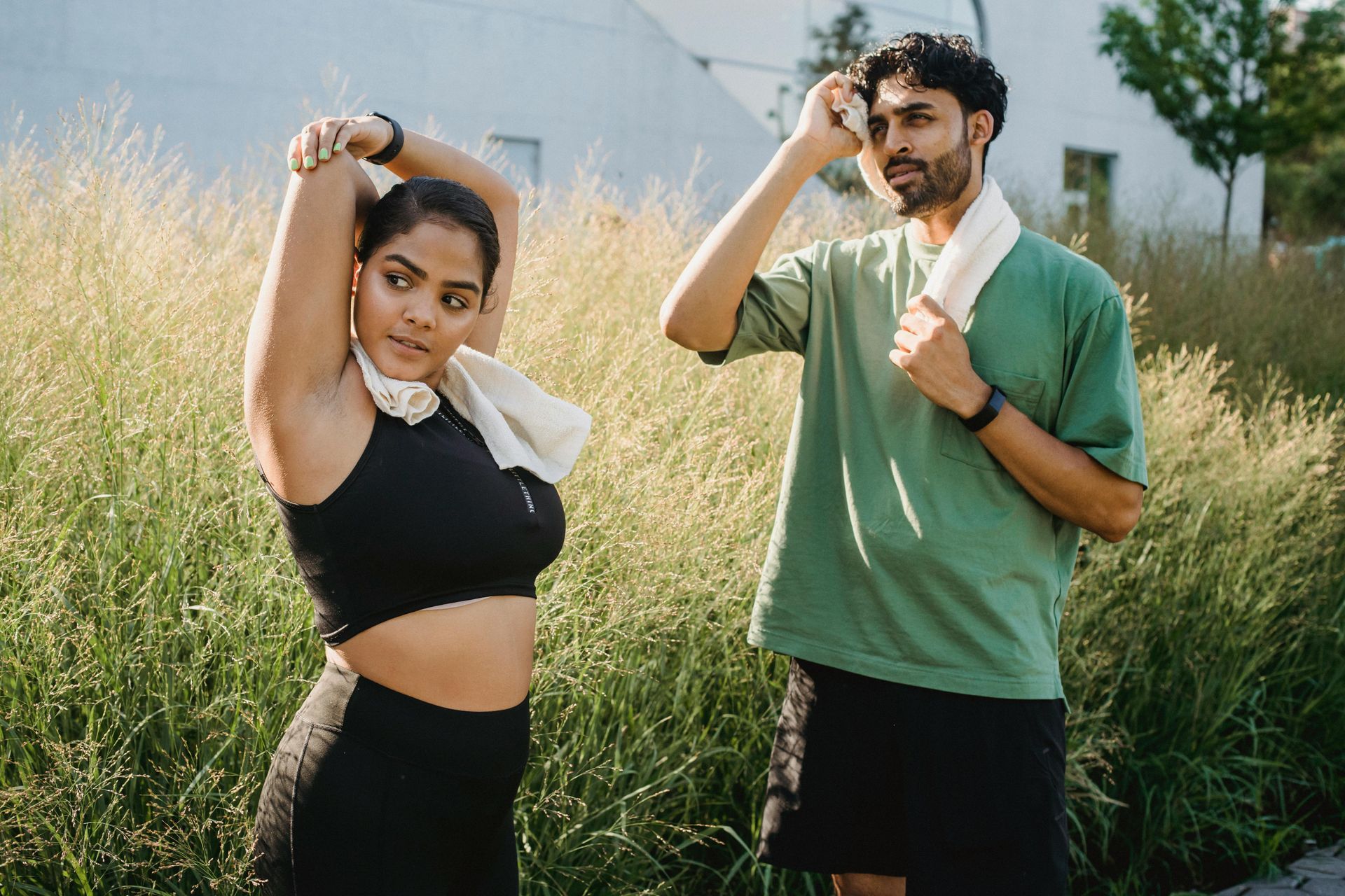 Woman stretching, man wiping sweat, both with towels, outdoors near green plants.