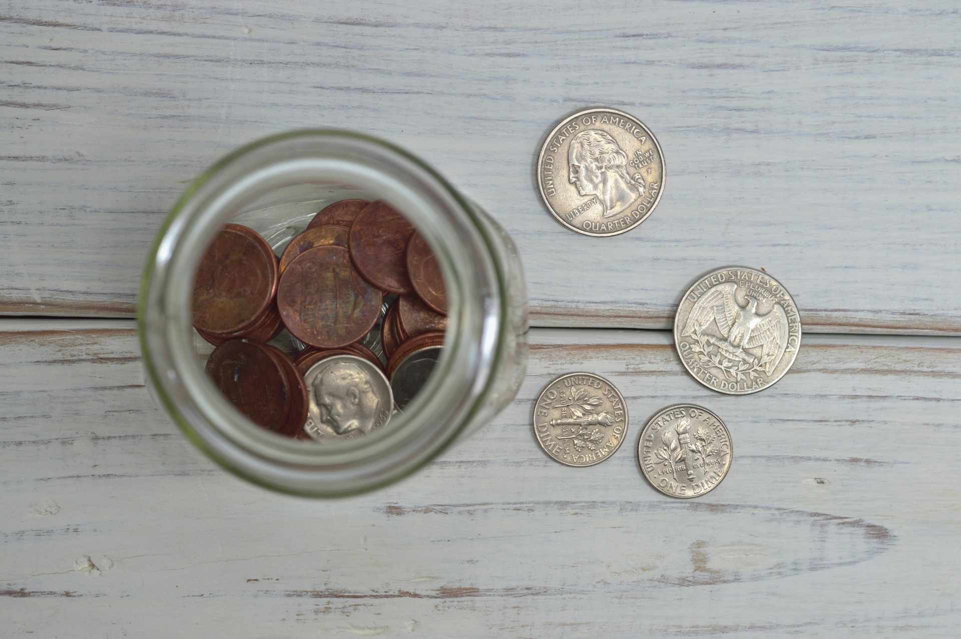 Glass jar with coins, spilling onto a light wood surface.