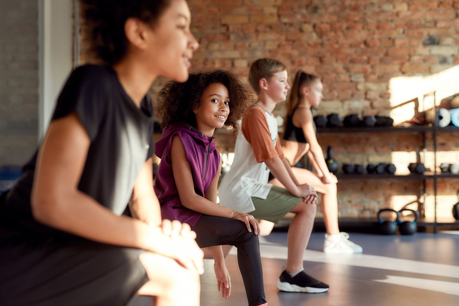 Four people doing squats in a gym. Smiling, they are near a brick wall and weights.