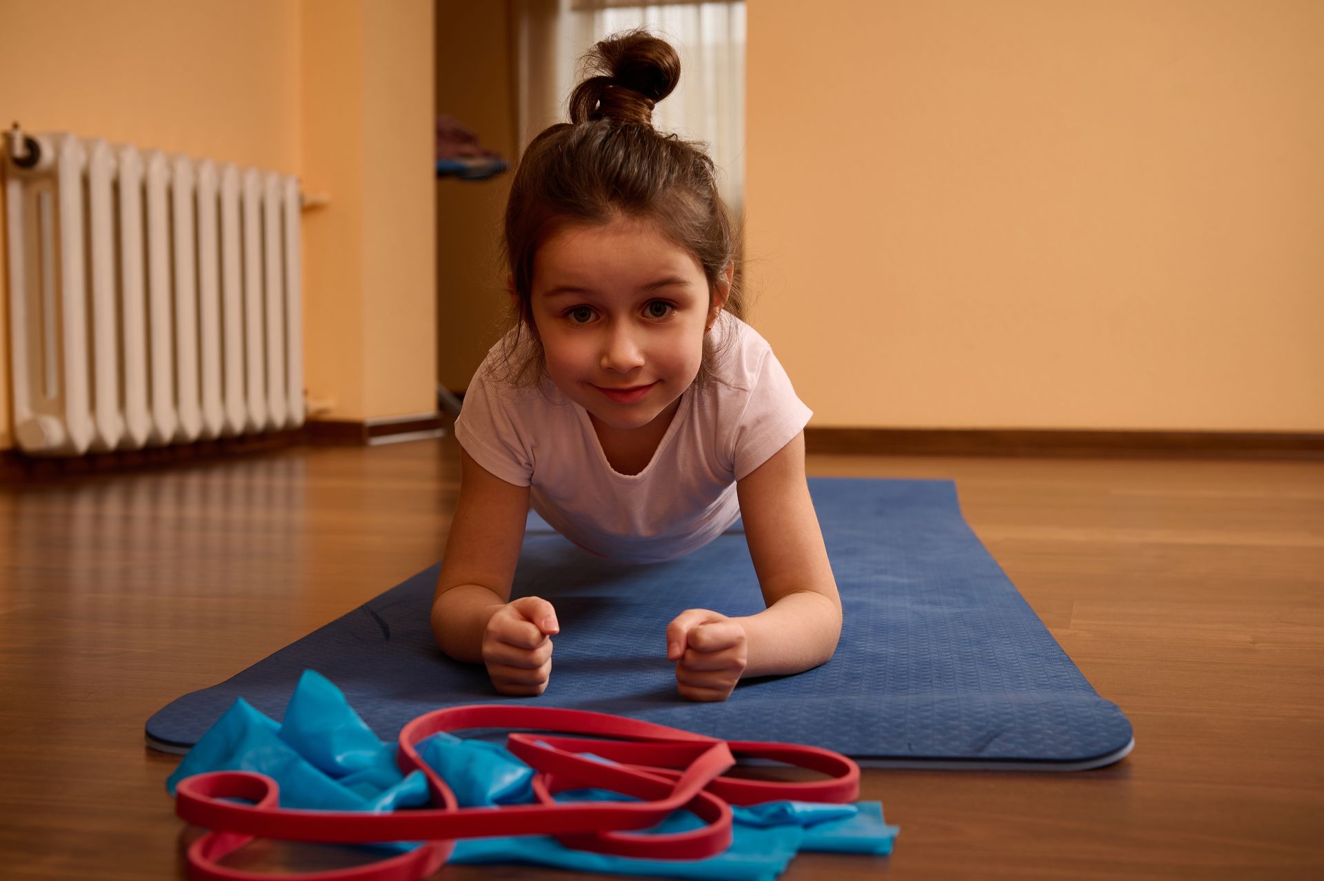 Girl doing a plank on a blue mat, looking at the camera. Resistance bands nearby. Indoors.