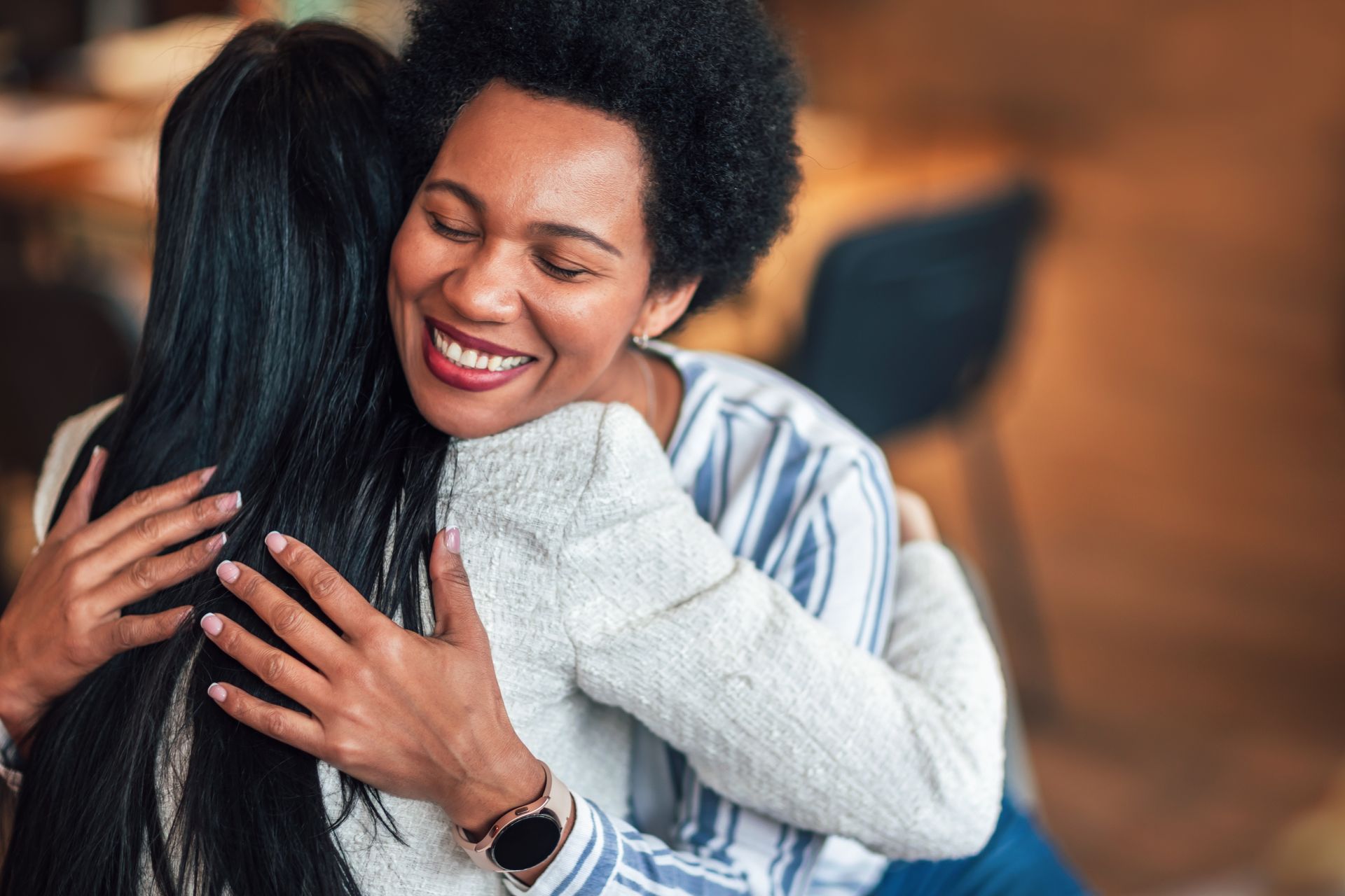 Two women embracing, smiling. One with dark hair and a watch, the other with short, curly hair. Blurred background.