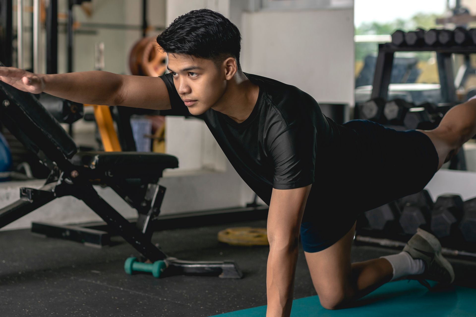 Man in gym doing bird-dog exercise, one arm and opposite leg extended. Gym setting, black shirt, blue mat.