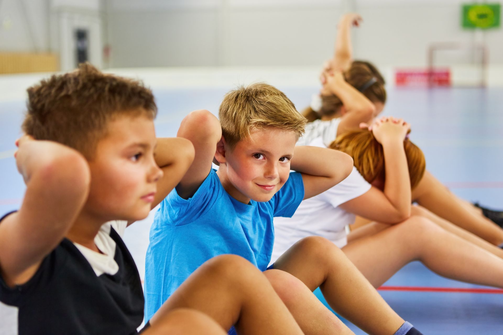 Children doing sit-ups on a gym floor, hands behind heads, one smiling.