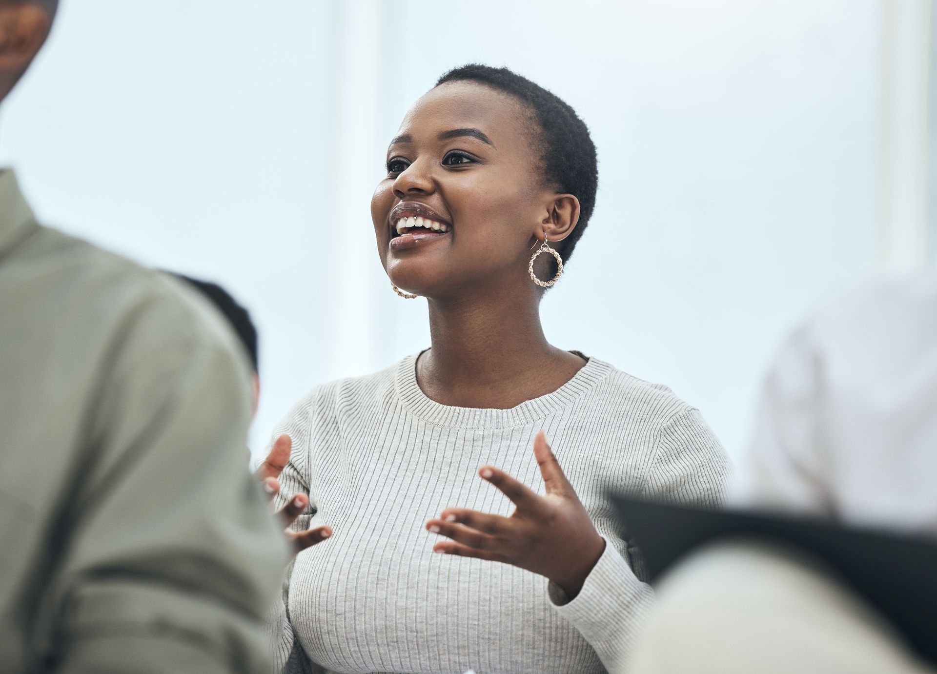 Woman speaking, gesturing with hands, smiling. Wearing a white sweater, hoop earrings.