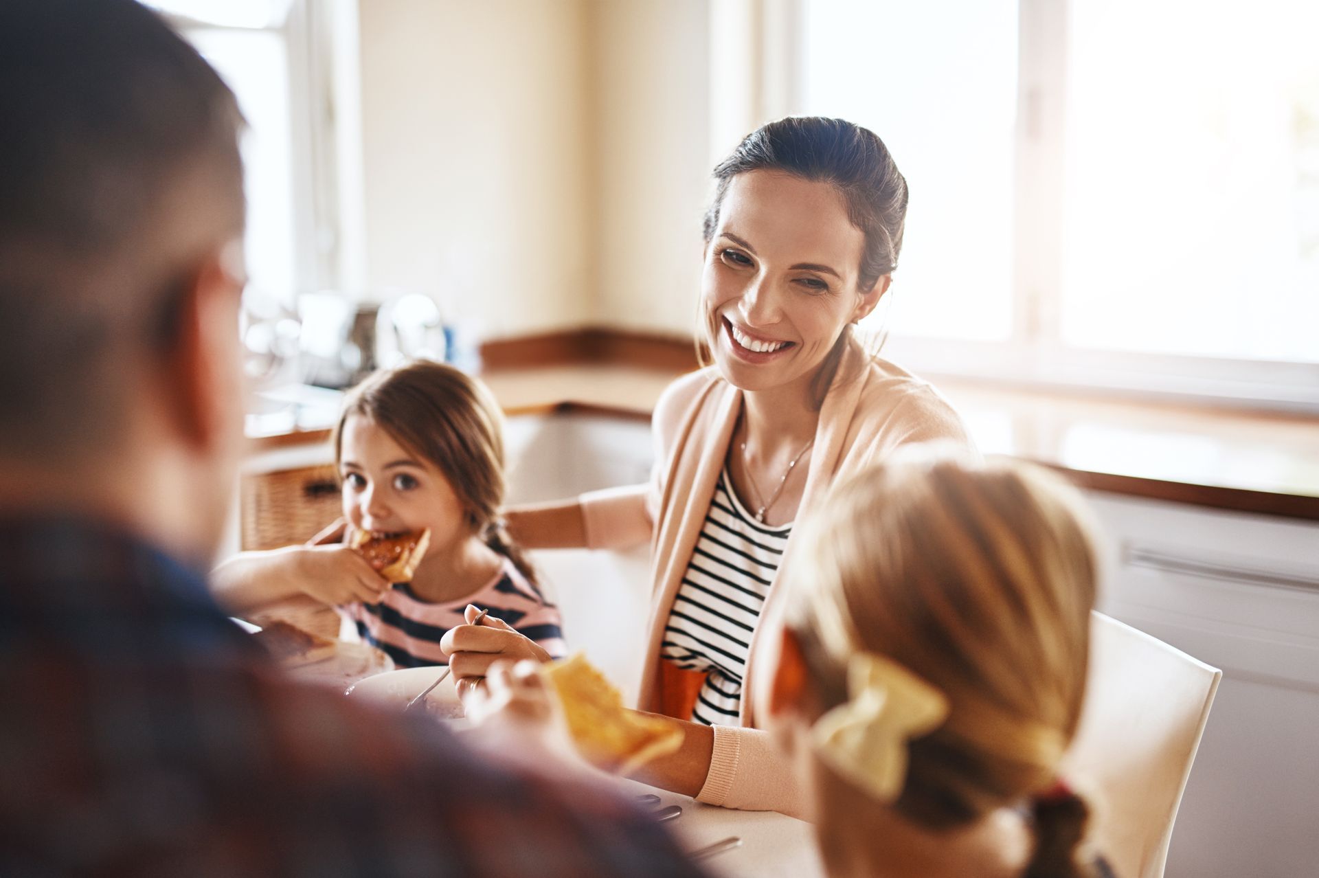 Family eating breakfast at a table; mother smiles at a child, sunlight streams in the window.