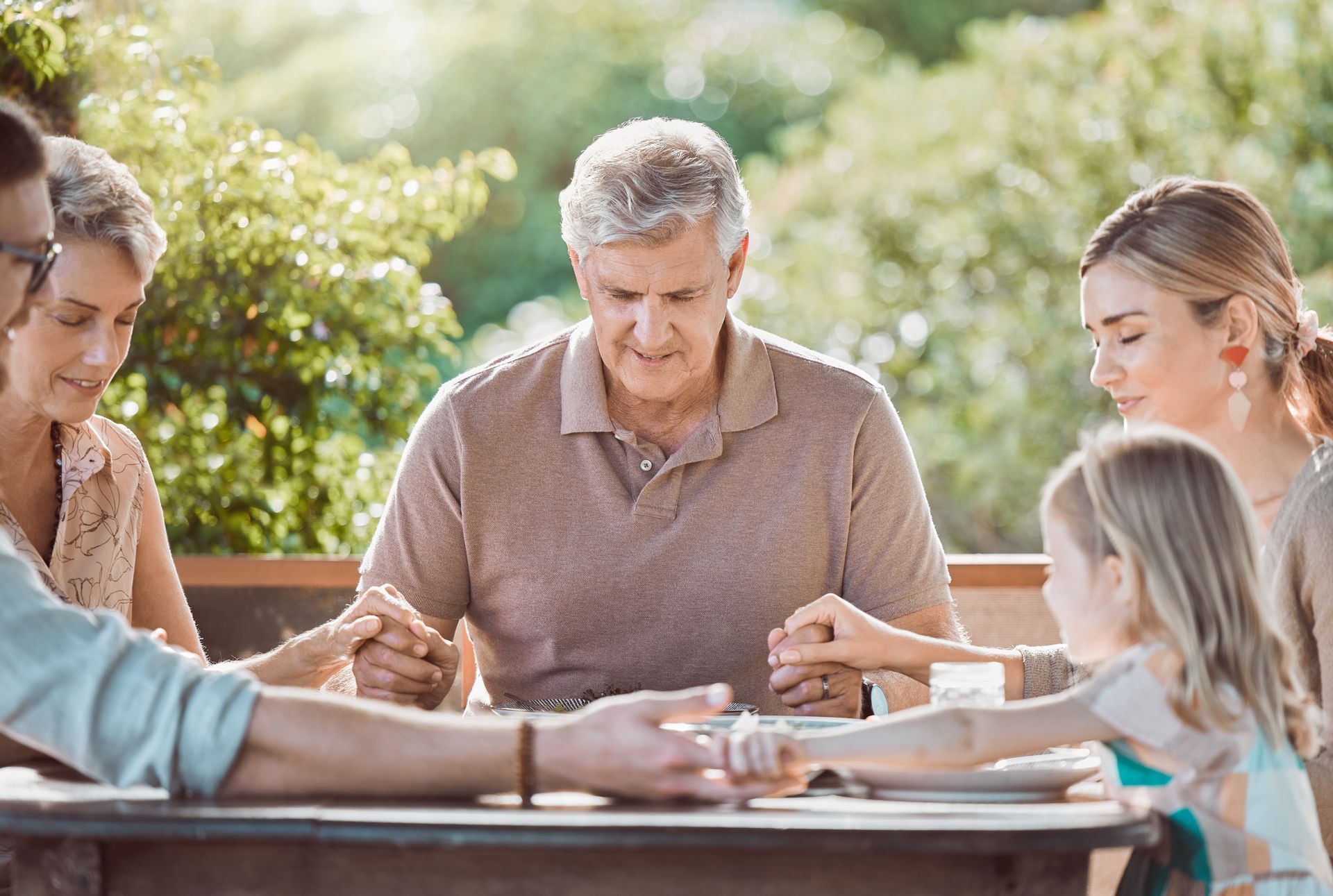 Family praying outdoors, holding hands at a table. Sunlight streams in.