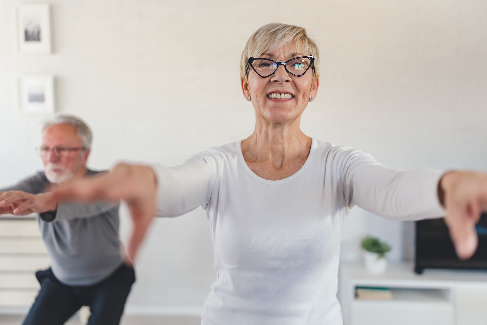 Woman with glasses and outstretched arms exercising with a man in the background.