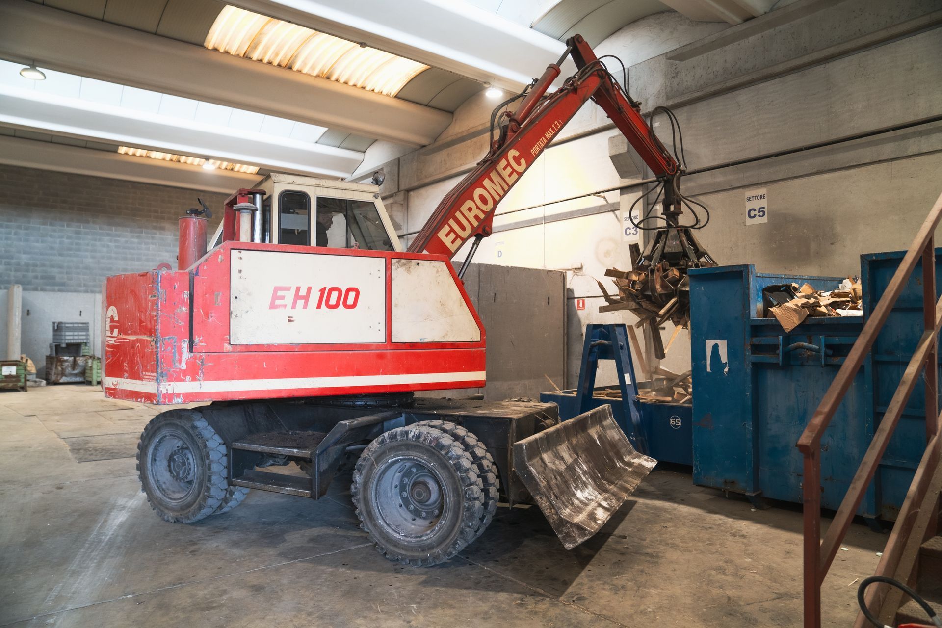 Escavatore EuroMec EH100 rosso e bianco in un magazzino che preleva rottami metallici 