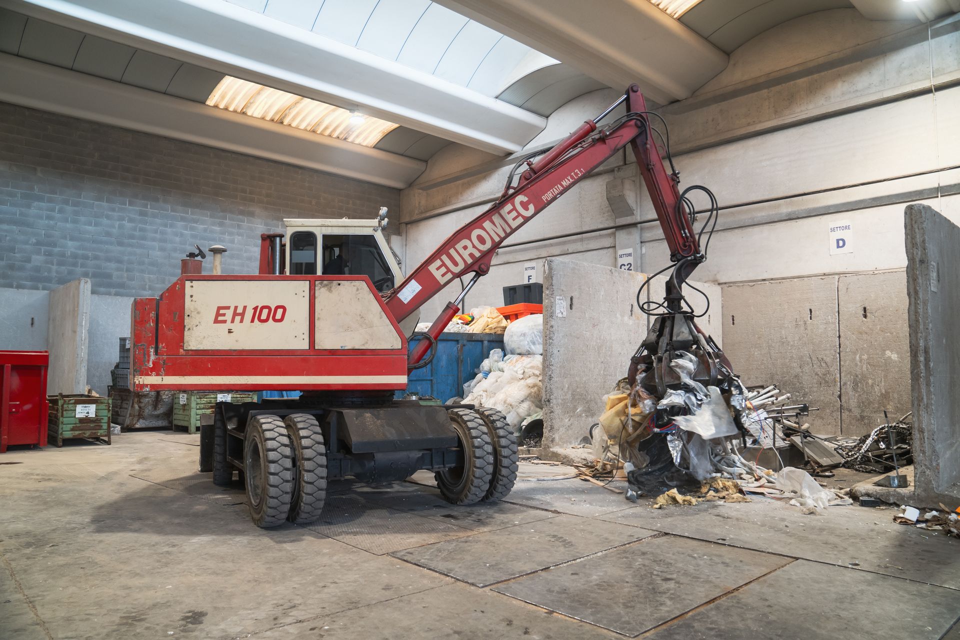 Macchina EUROMEC EH100 rossa e bianca che afferra e solleva rottami metallici in un impianto di riciclaggio.