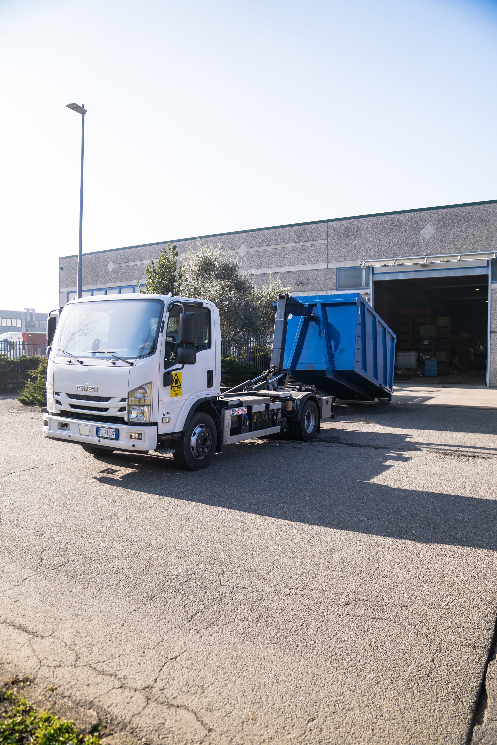 White truck with a blue dumpster on a gravel lot in front of a building.