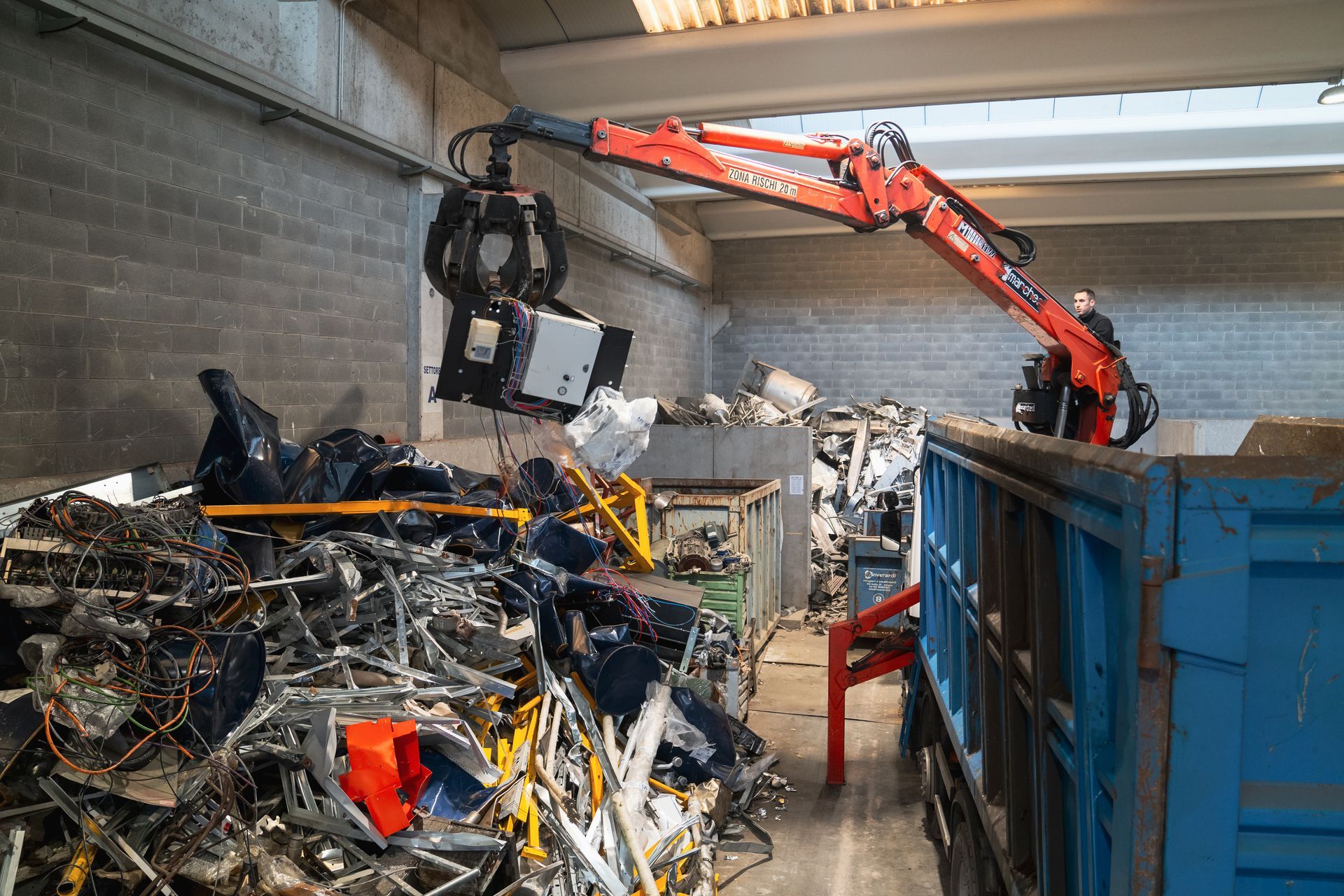 Una gru solleva del metallo da una pila e lo carica in un contenitore blu in un impianto di riciclaggio.