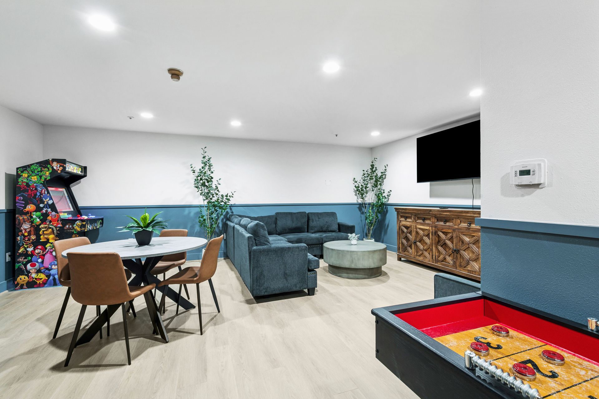 Living room with arcade game, sofa, table, and shuffleboard. White and blue walls with wooden accents.