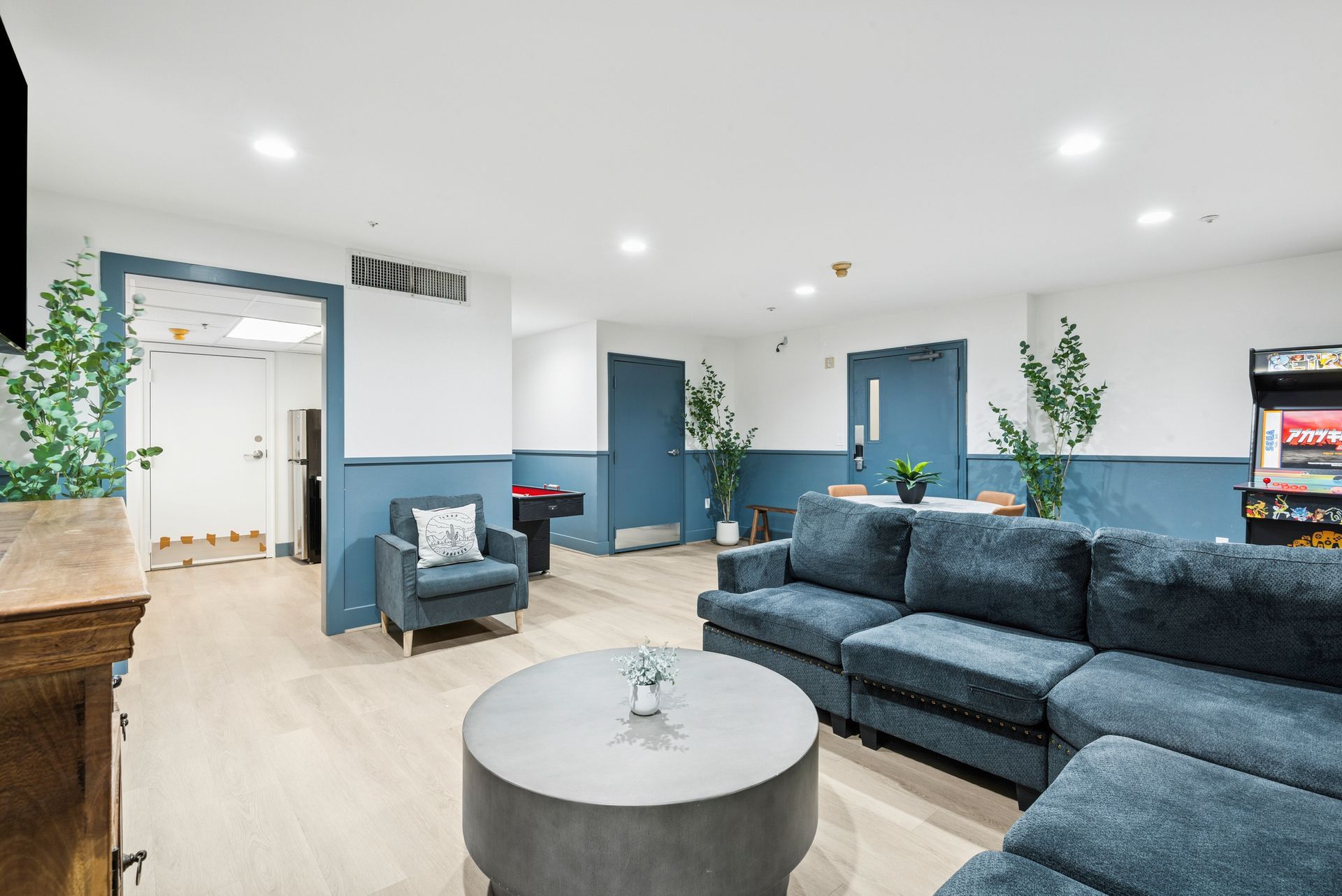 Lounge with blue sectional sofa, armchair, and round coffee table. Arcade game, plants, and doors in the room.