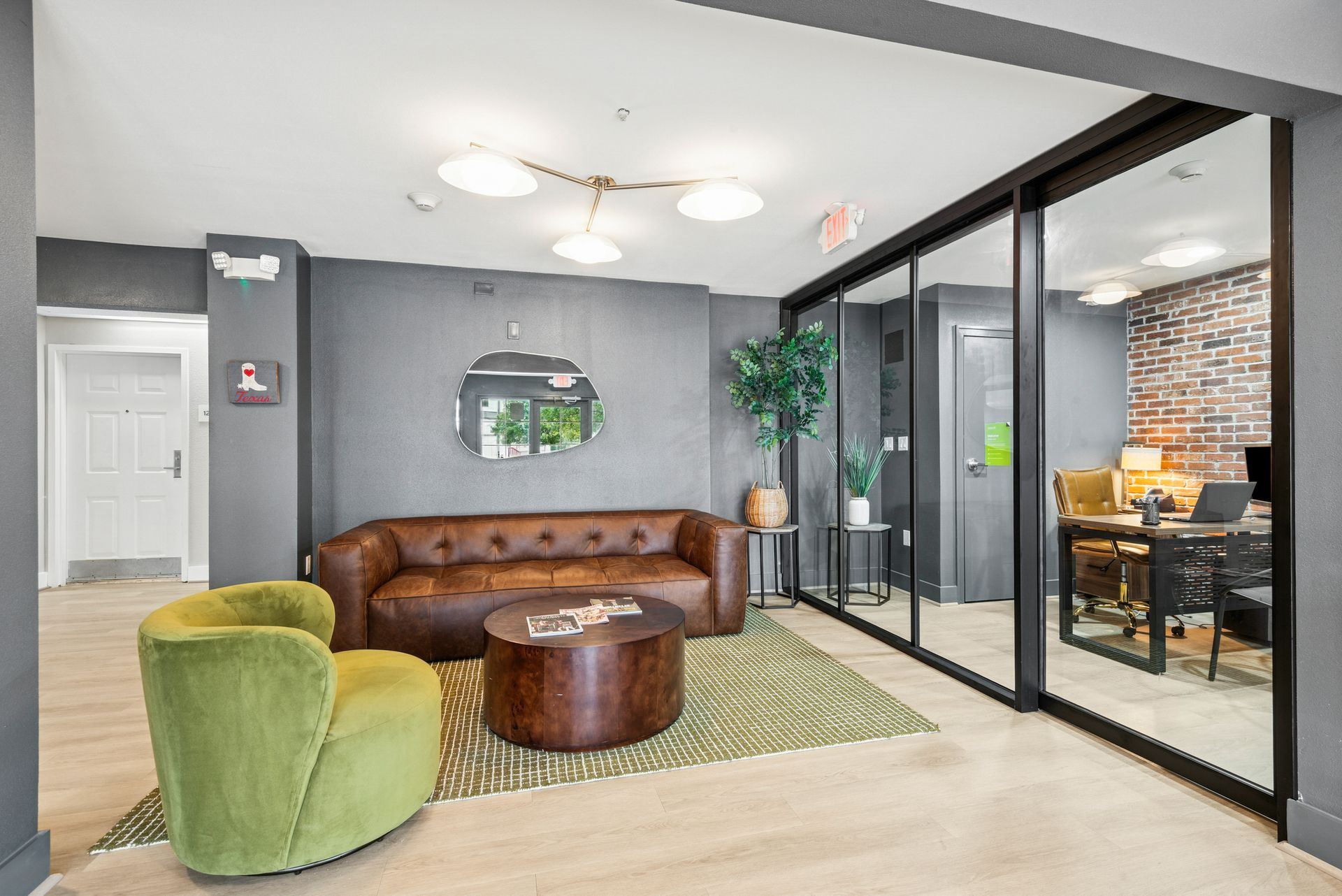 Cozy lounge with brown sofa, green chair, round coffee table, rug, glass wall to a work area.