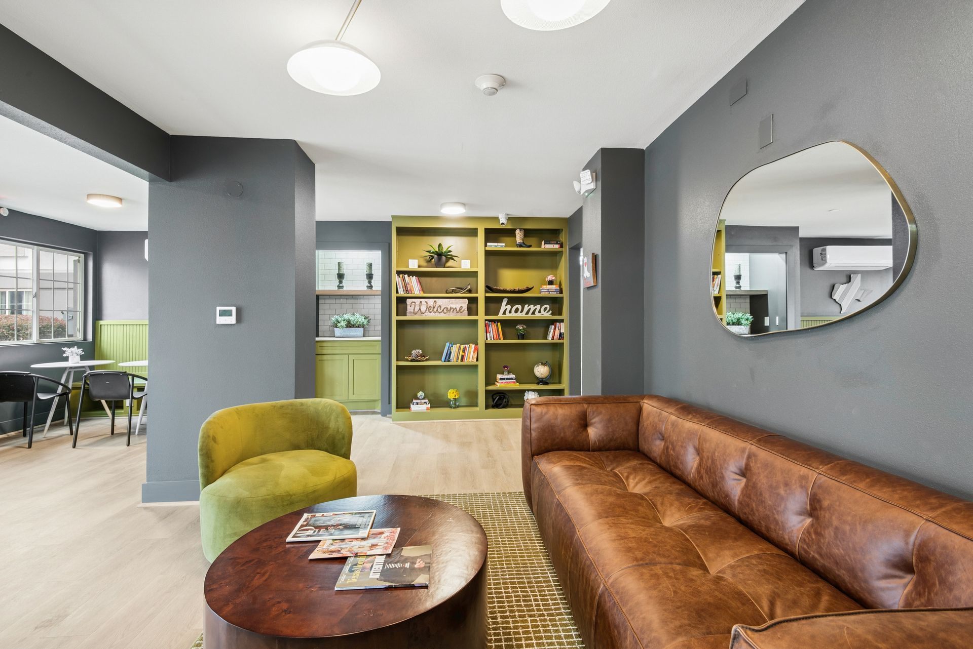 Lounge area with brown leather sofa, green armchair, and round coffee table. Gray walls and olive green bookshelf.