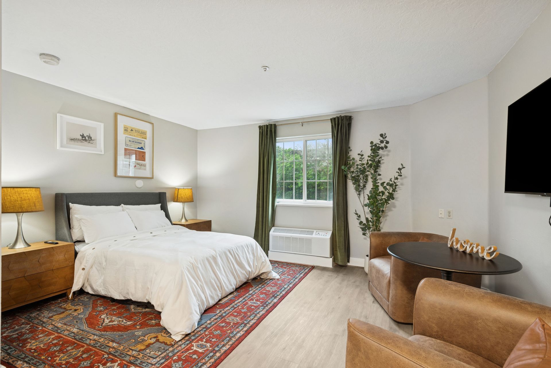 Cozy studio apartment with a bed, small table, and TV on the wall. Light gray walls, red rug, and green curtains.