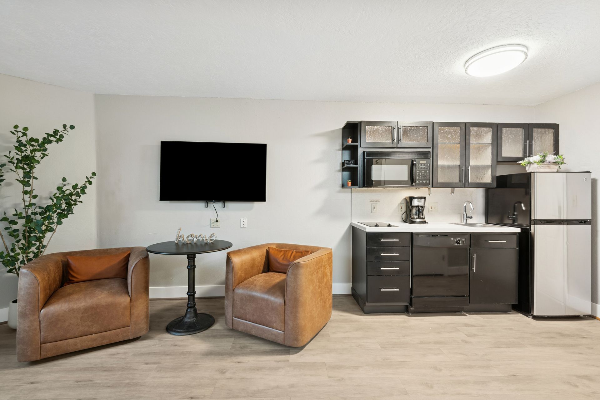 Cozy studio apartment with brown leather armchairs, small kitchen, and a mounted TV.