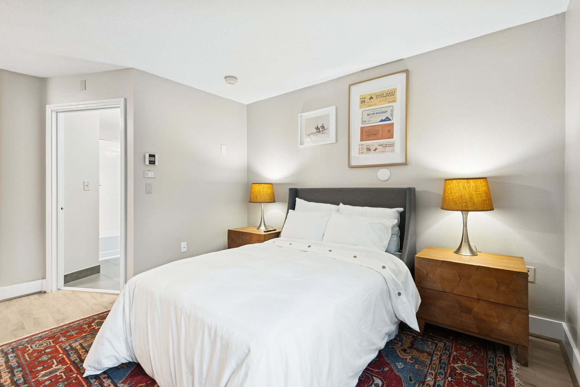 Bedroom with bed, nightstands, lamps, artwork, and area rug. A door leads to another room.