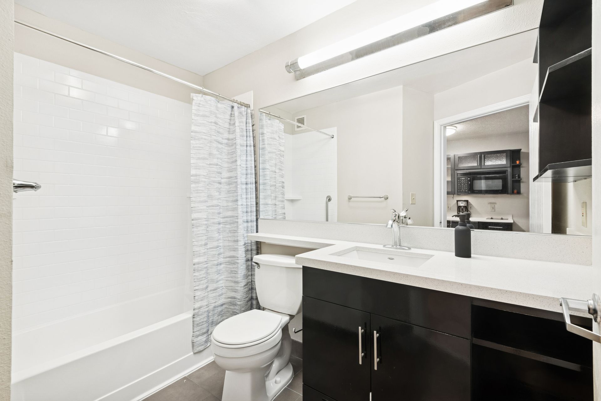 Bathroom with white tub/shower, toilet, dark cabinet, large mirror, and open doorway.