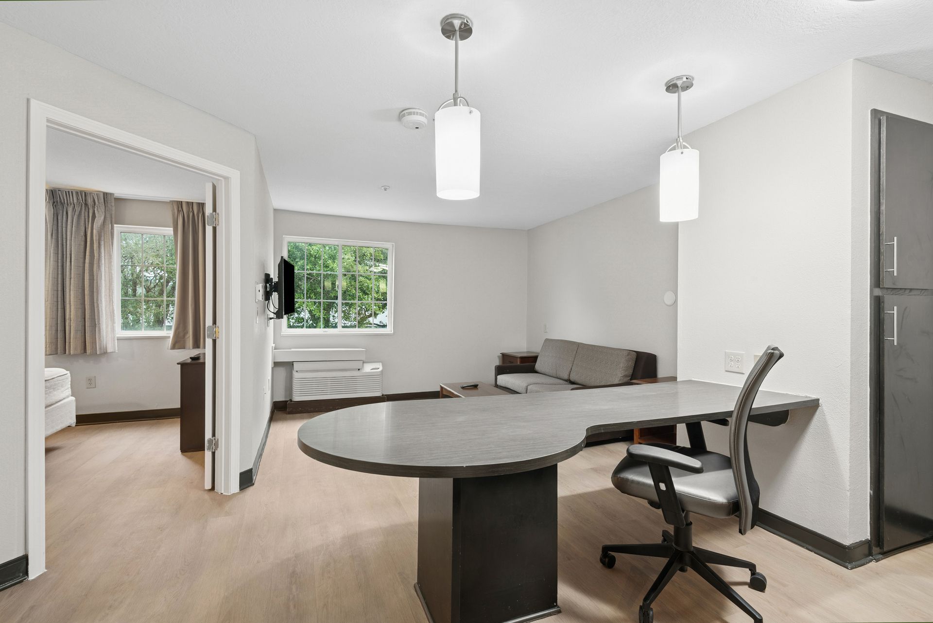 Living room with desk, sofa, and a view through a window. Light-colored walls, wood floor.
