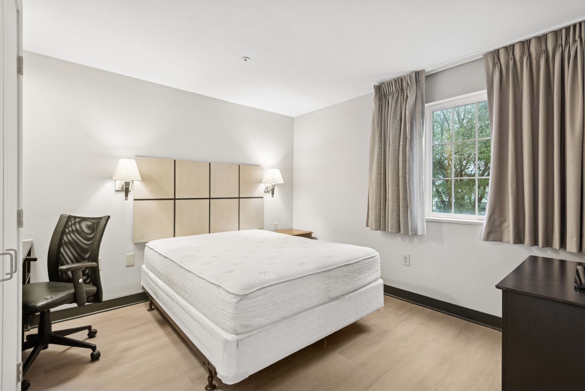 Bedroom with a bed, desk, chair, and window with curtains. Neutral colors and simple decor.