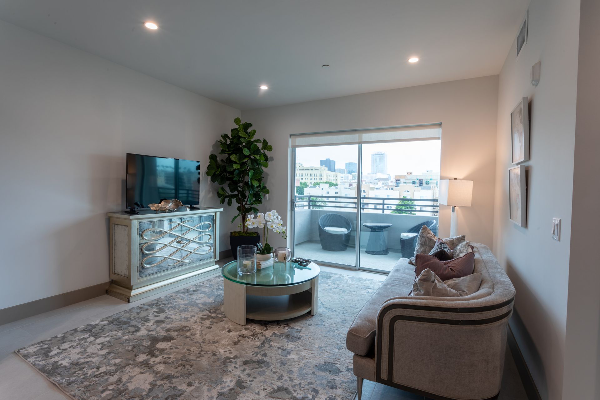 Living room with balcony, features sofa, mirrored dresser with TV, rug, and city view.