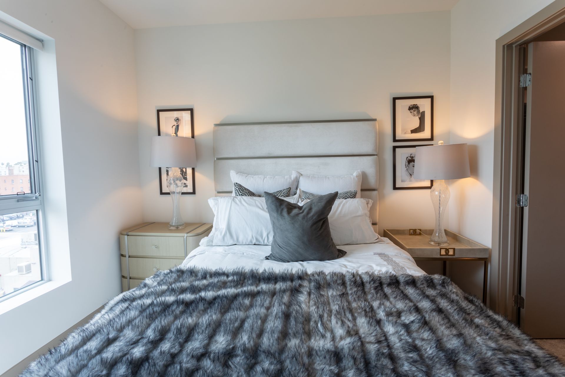 Bedroom with bed, nightstands, lamps, artwork, and window. Gray fur blanket and neutral color scheme.