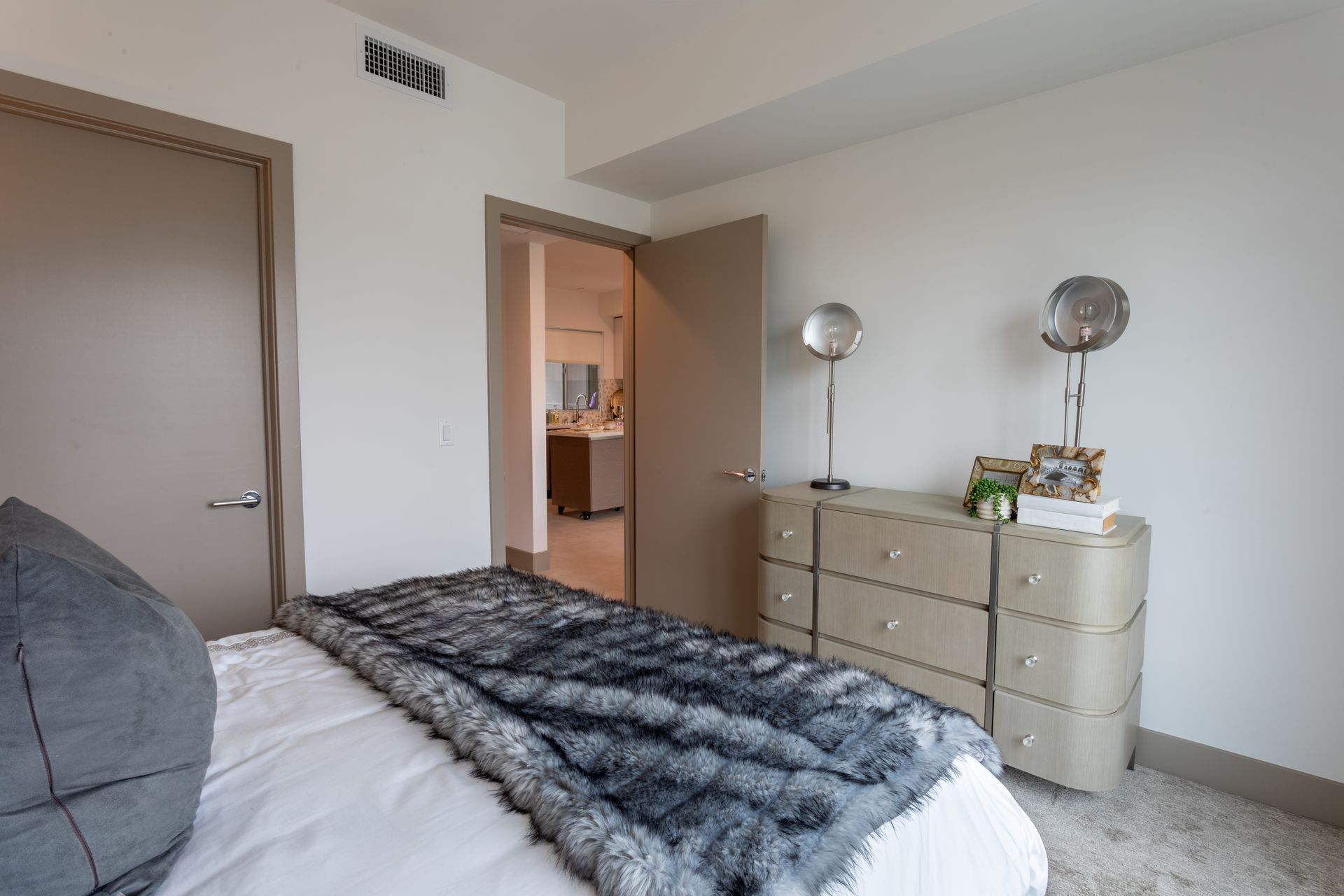 Bedroom with bed, dresser, doors, and a glimpse of a bathroom. Neutral colors.