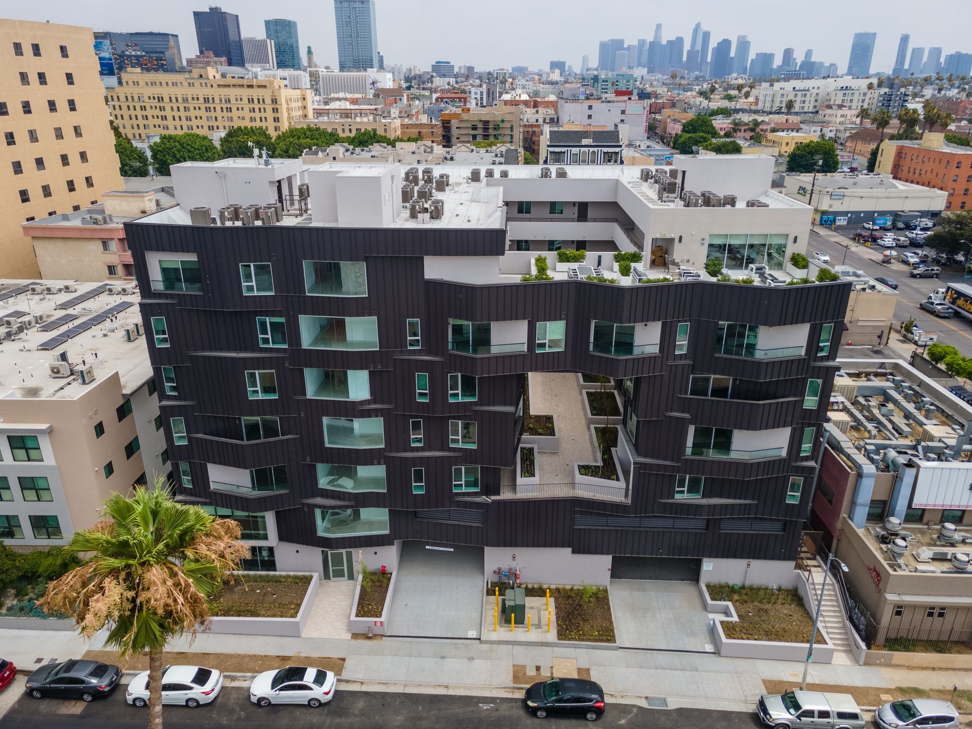 Modern black building with open courtyard, overlooking a cityscape. Palm tree in foreground, cars parked below.