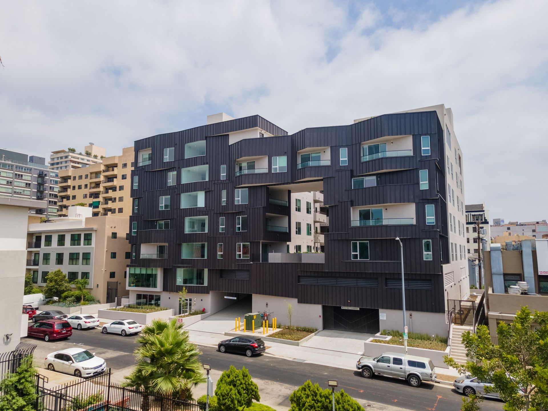 Modern, dark-clad apartment building with multiple windows. Street view with cars parked and driving. Blue sky.