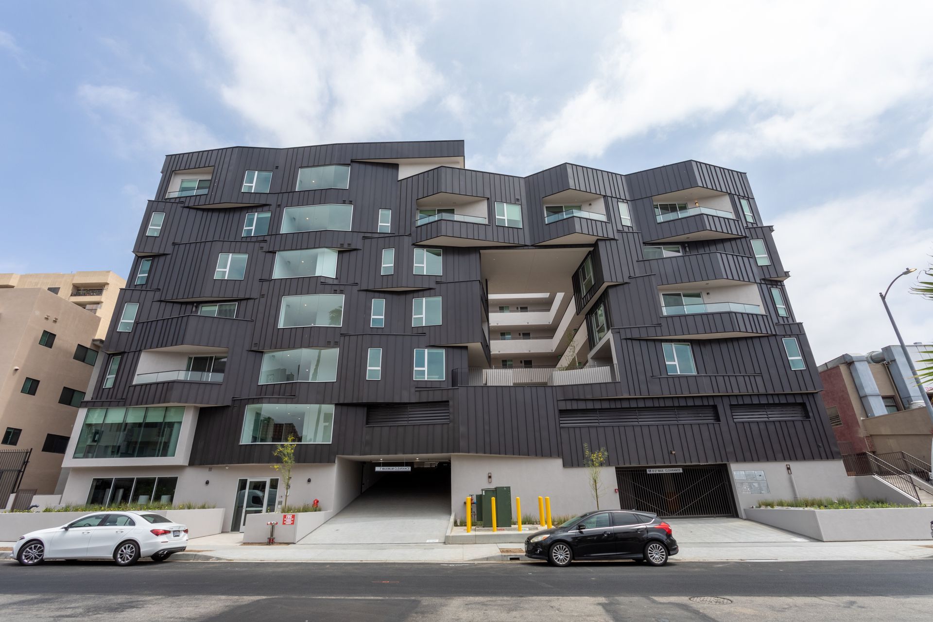 Modern black apartment building with irregularly shaped facade, parking garage, and cars parked on street.