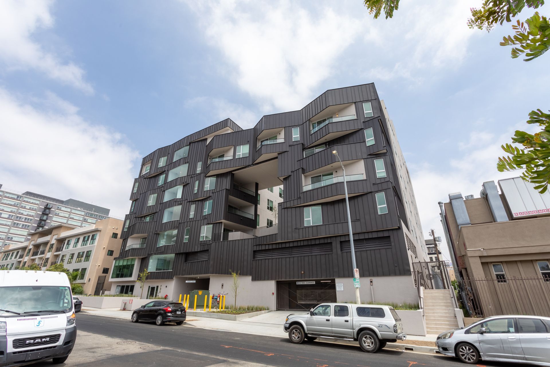 Black modern apartment building on a street with parked cars and a utility pole, under a partly cloudy sky.