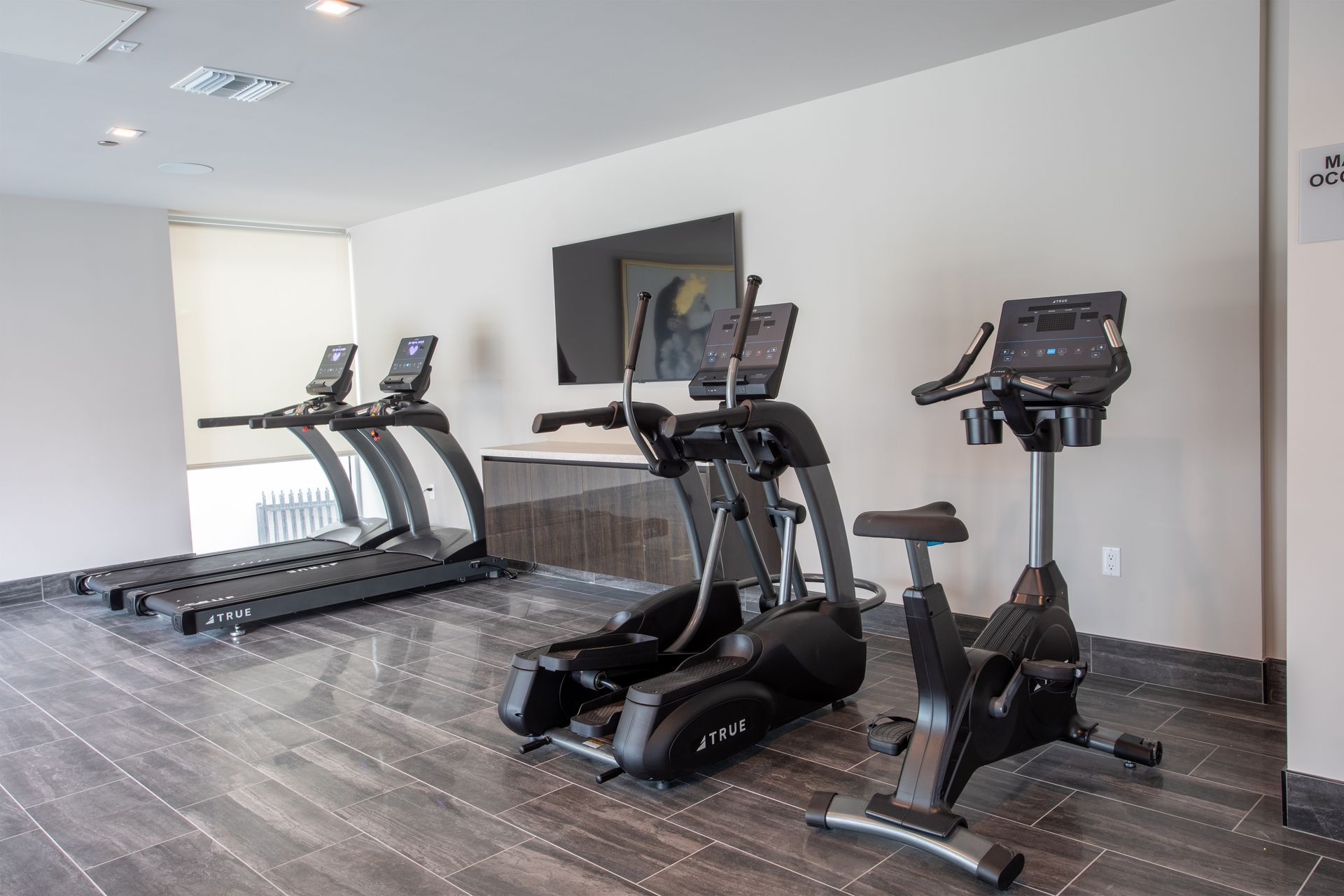 Gym interior with treadmills, elliptical, stationary bike, and TV against a light wall.