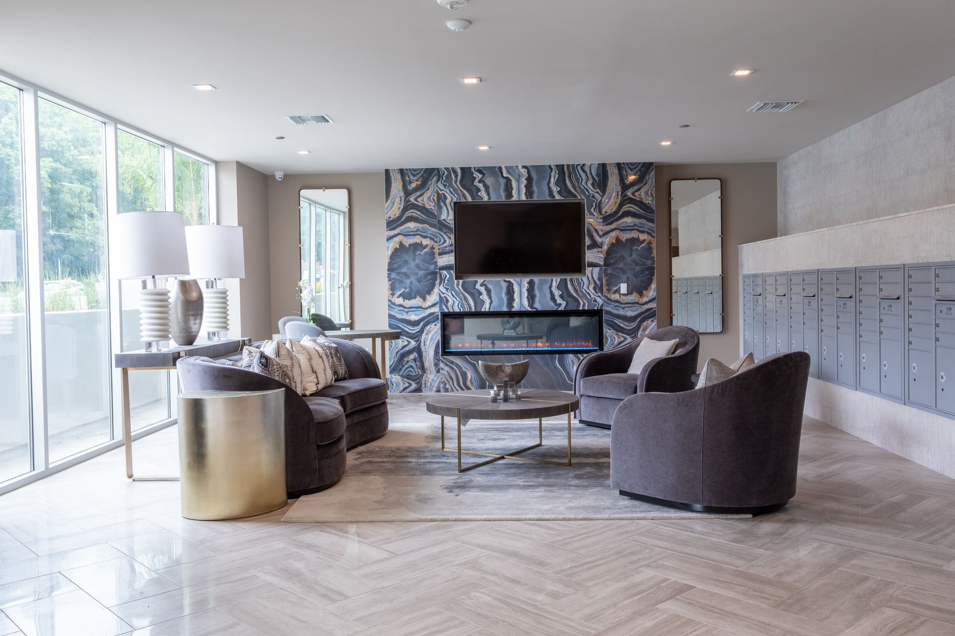 Lobby with seating, fireplace, and mailboxes. Gray armchairs, a rug, and a gold side table. Large windows.