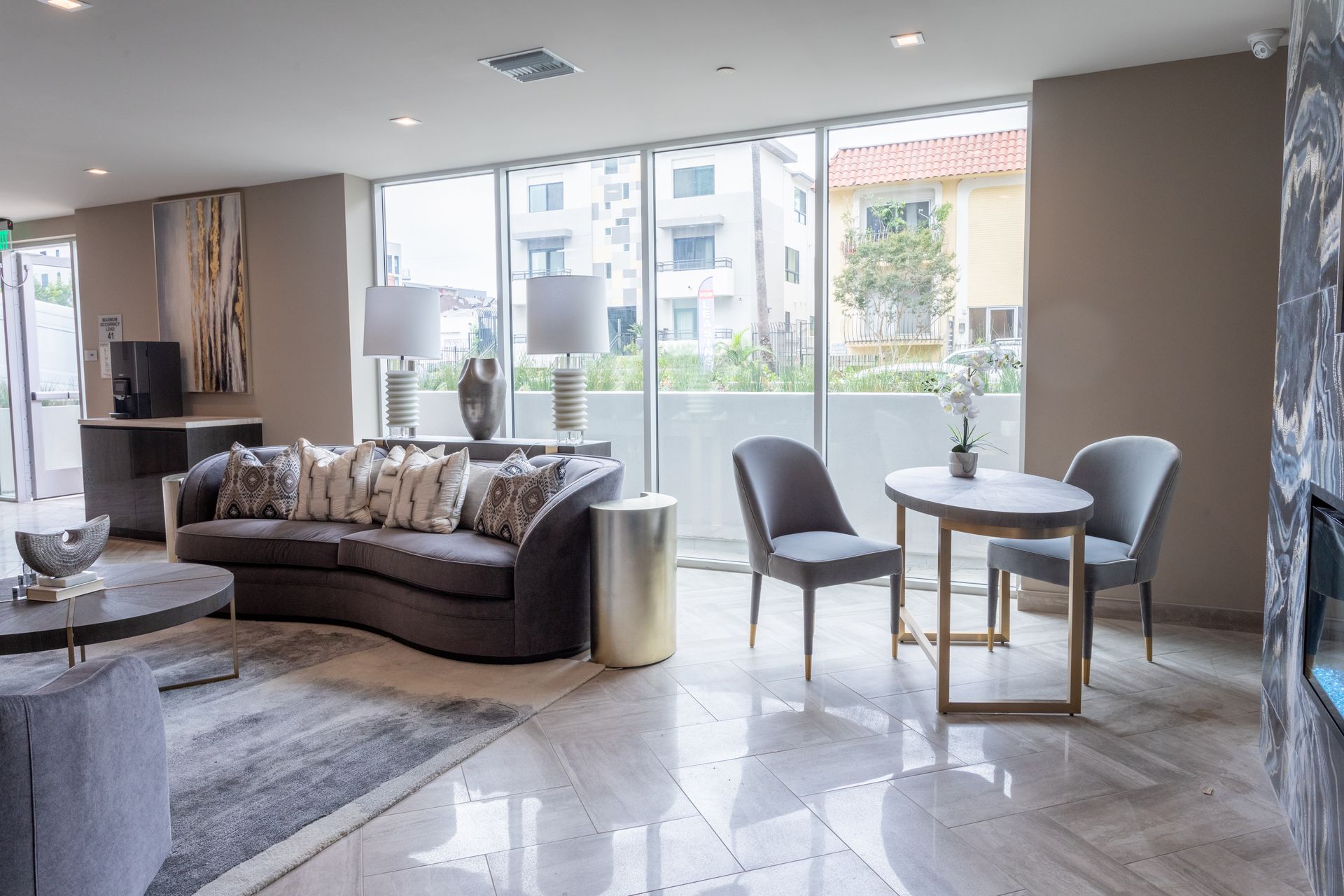 Modern lobby with gray couch, small table, chairs, large windows, and marble floors.