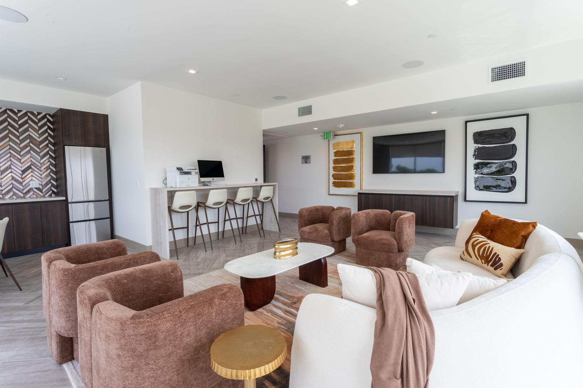 Modern lounge with seating, a bar, a TV, and a kitchenette. Neutral colors, warm accents.